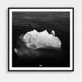 A framed black and white photographic print of an iceberg floating in dark water, presented in a square black frame with a white mat against a light gray wall.