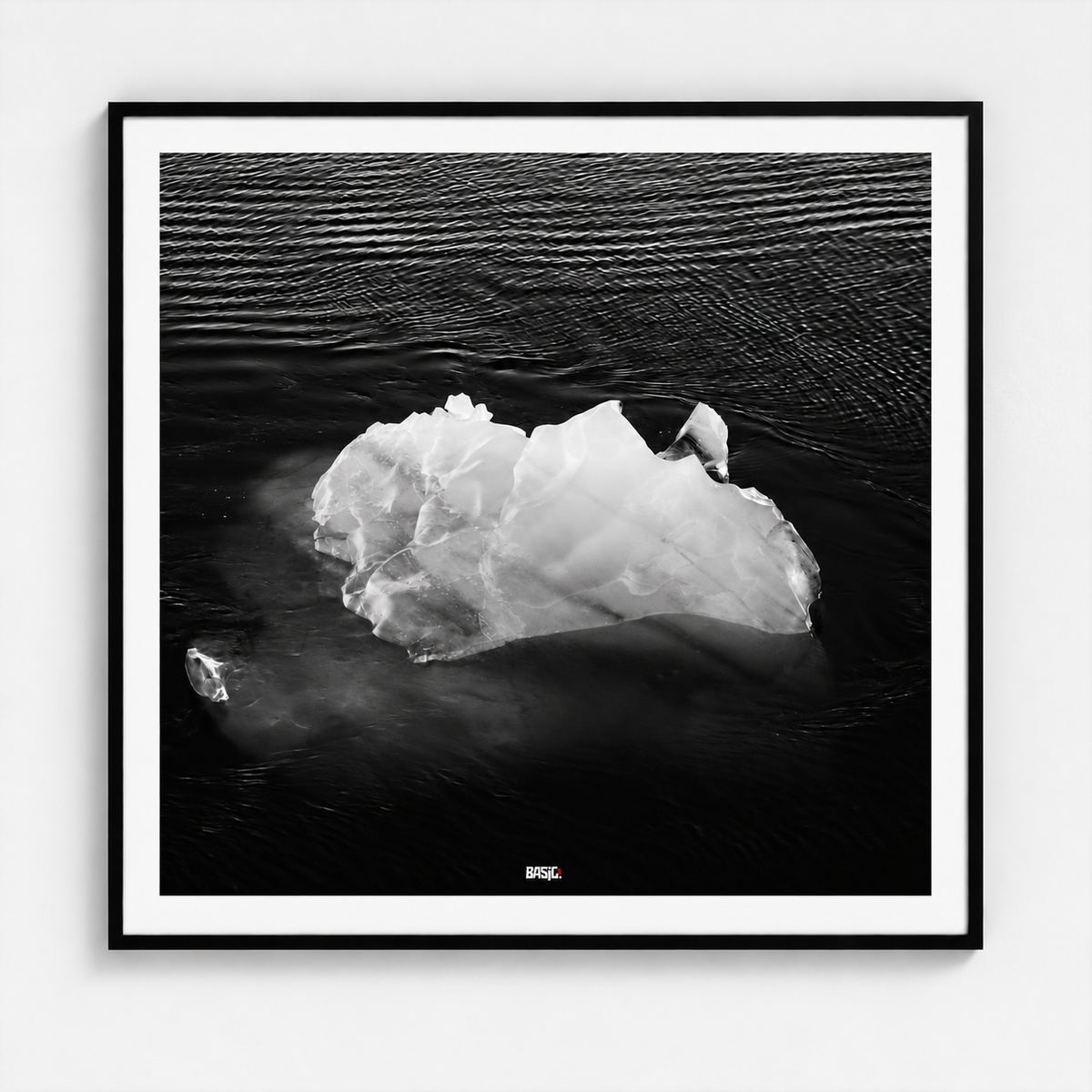 A framed black and white photographic print of an iceberg floating in dark water, presented in a square black frame with a white mat against a light gray wall.