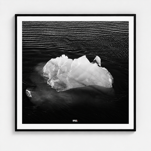 A framed black and white photographic print of an iceberg floating in dark water, presented in a square black frame with a white mat against a light gray wall.