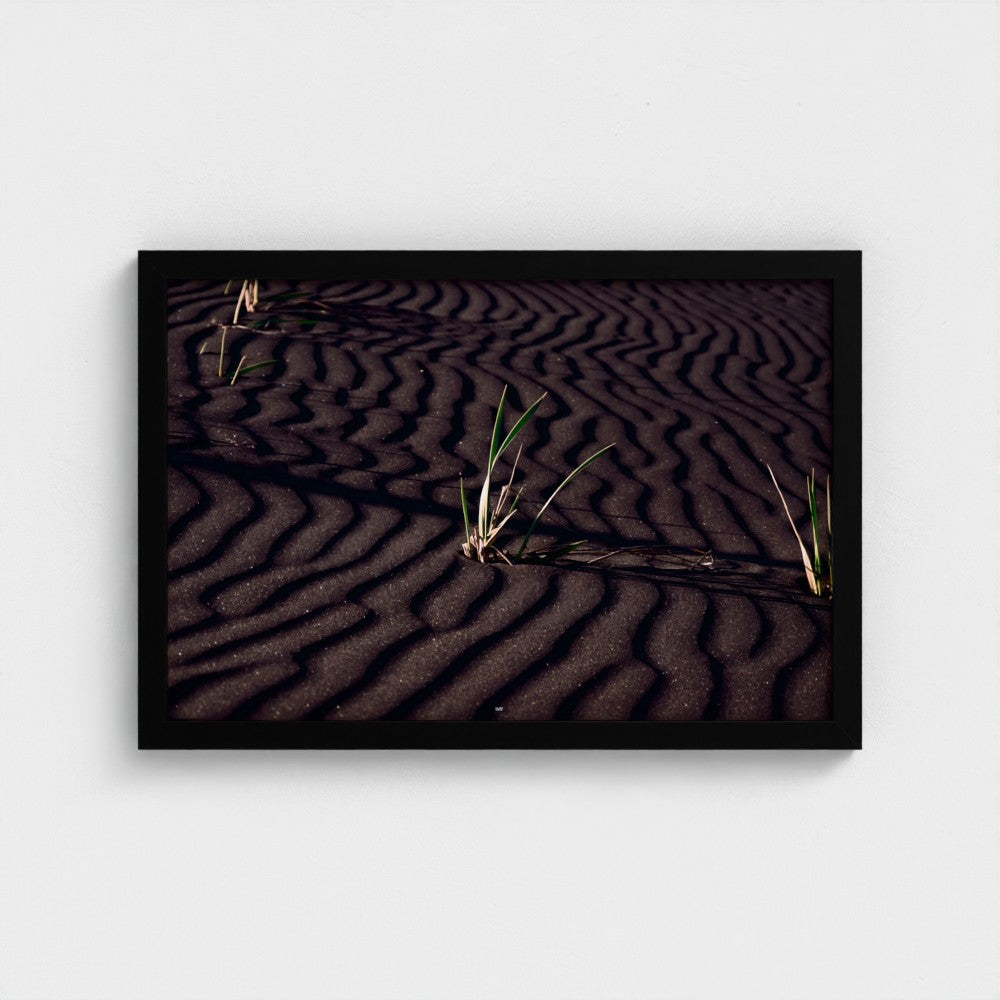 A framed square art print depicting dark rippled sand dunes with small green grasses, presented in a black frame against a light gray wall.