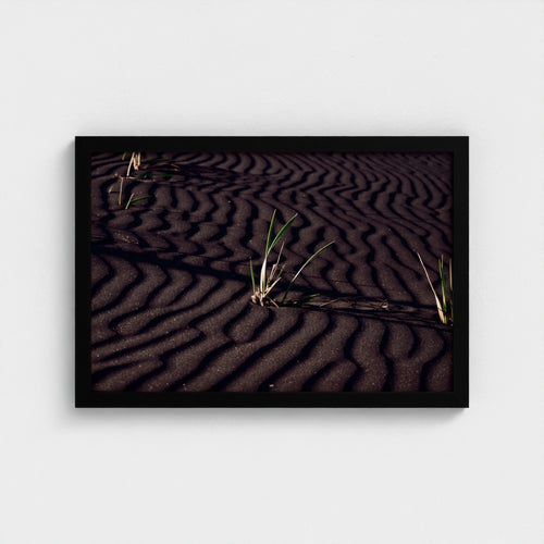 A framed square art print depicting dark rippled sand dunes with small green grasses, presented in a black frame against a light gray wall.