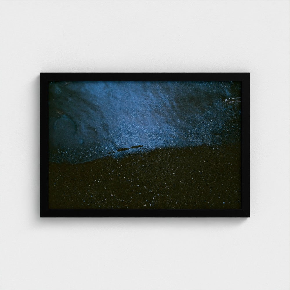 A framed abstract art print featuring a textured blue and dark brown composition, housed in a simple black frame and mounted on a light gray wall.