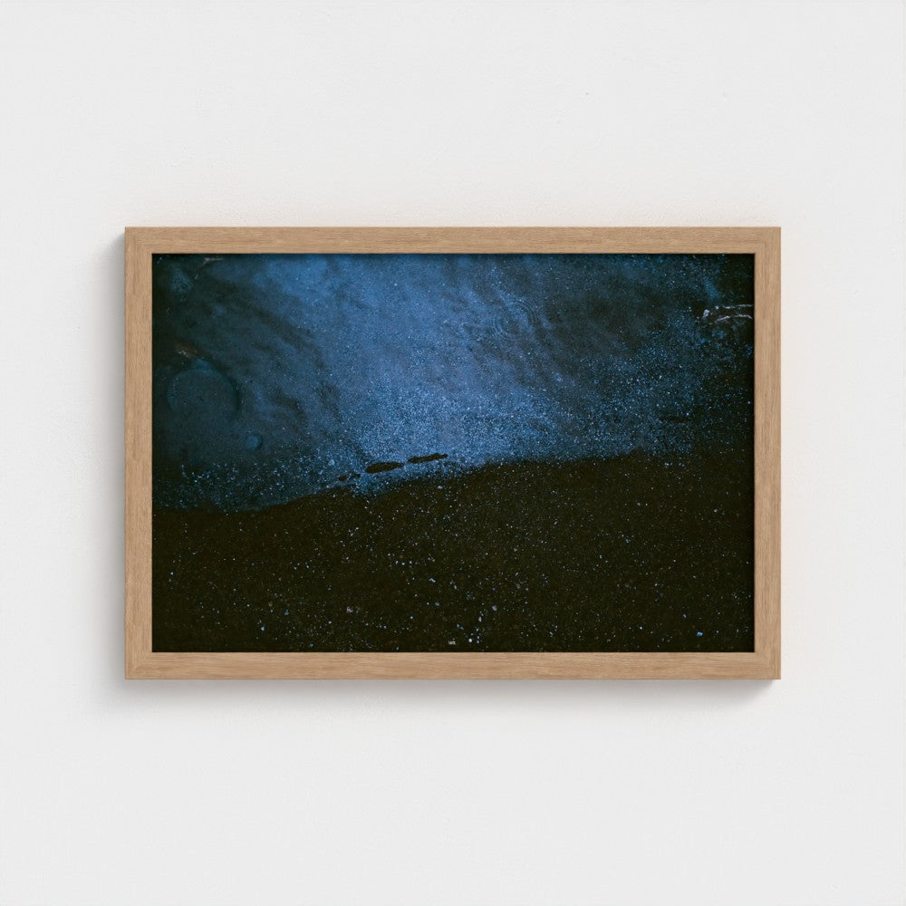 A framed abstract art print featuring a textured dark blue and black landscape, housed in a light natural wood frame and mounted on a clean white wall.