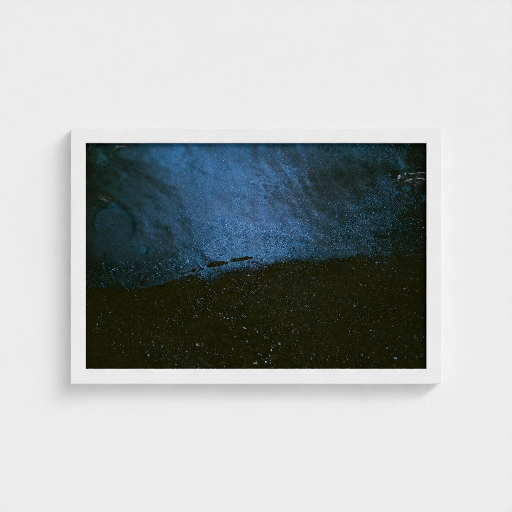 A framed abstract art print featuring a textured, fluid composition of deep blue and black tones, displayed against a clean white wall.