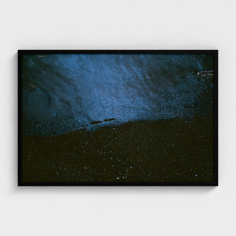 A framed abstract art print featuring a textured composition of deep blue and black tones with fine speckled details, presented in a black frame against a light gray wall.
