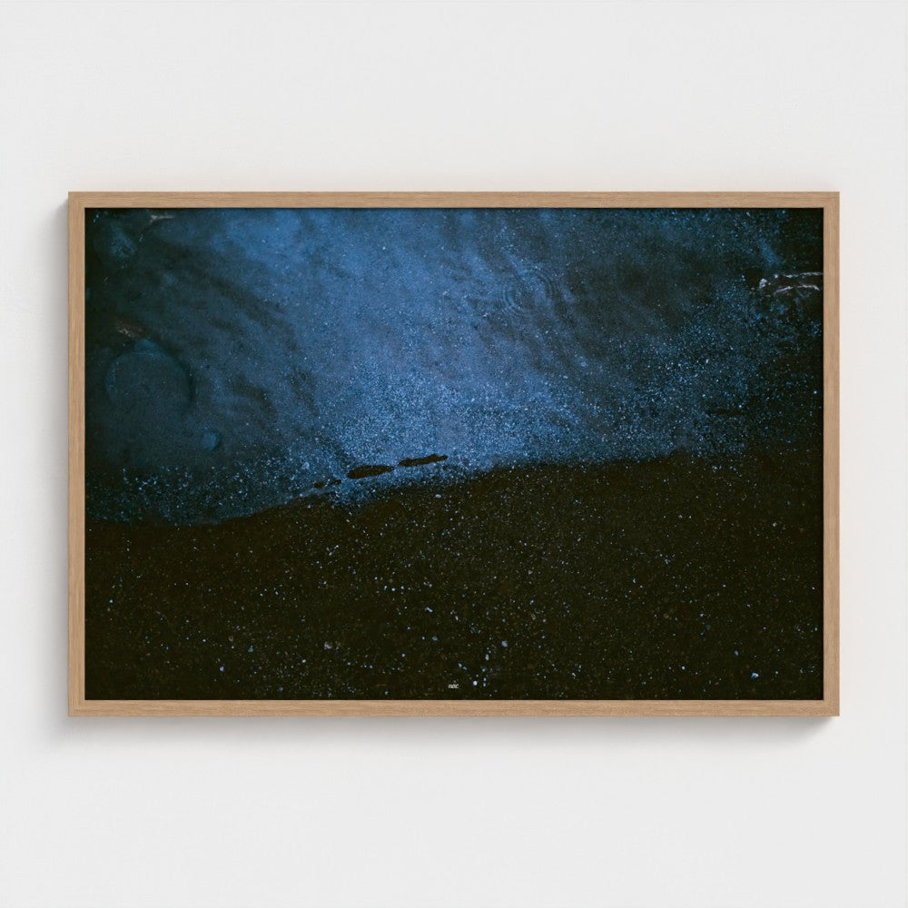 A framed abstract art print featuring a textured blue and black composition, housed in a light natural wood frame and mounted on a clean white wall.