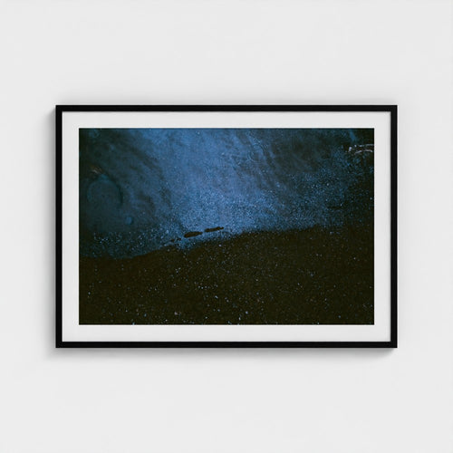 A framed abstract art print featuring a textured composition of deep blue and black tones, presented in a black frame with a white mat against a light gray wall.