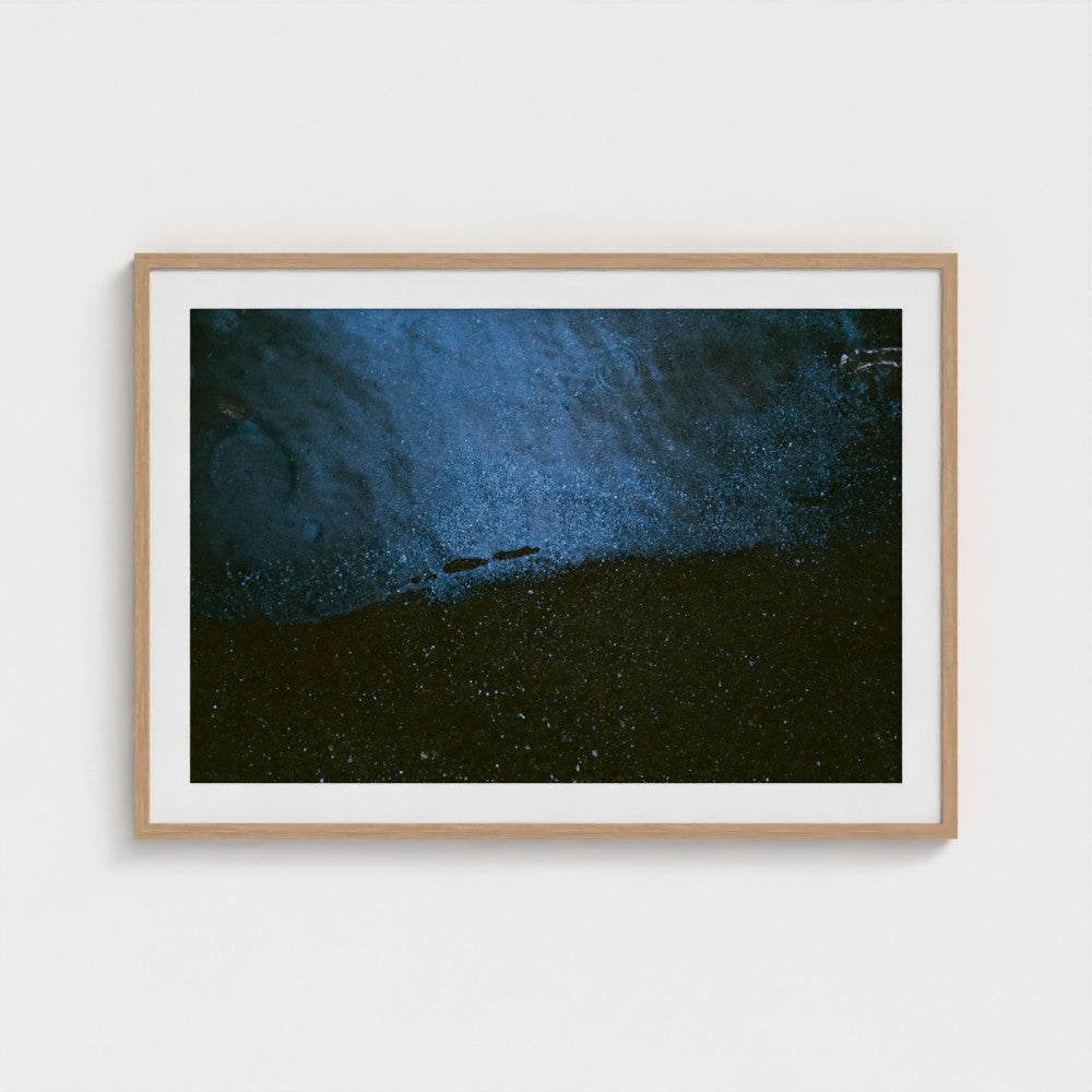 A framed art print featuring a dark, textured scene with deep blue and black tones, presented in a light natural wood frame against a clean white wall.