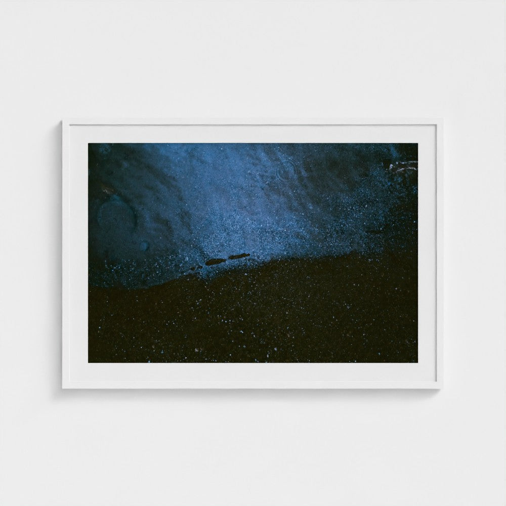 A framed abstract art print featuring a dark blue and black textured composition with a small horizontal bar element near the center, displayed in a white frame against a light gray wall.