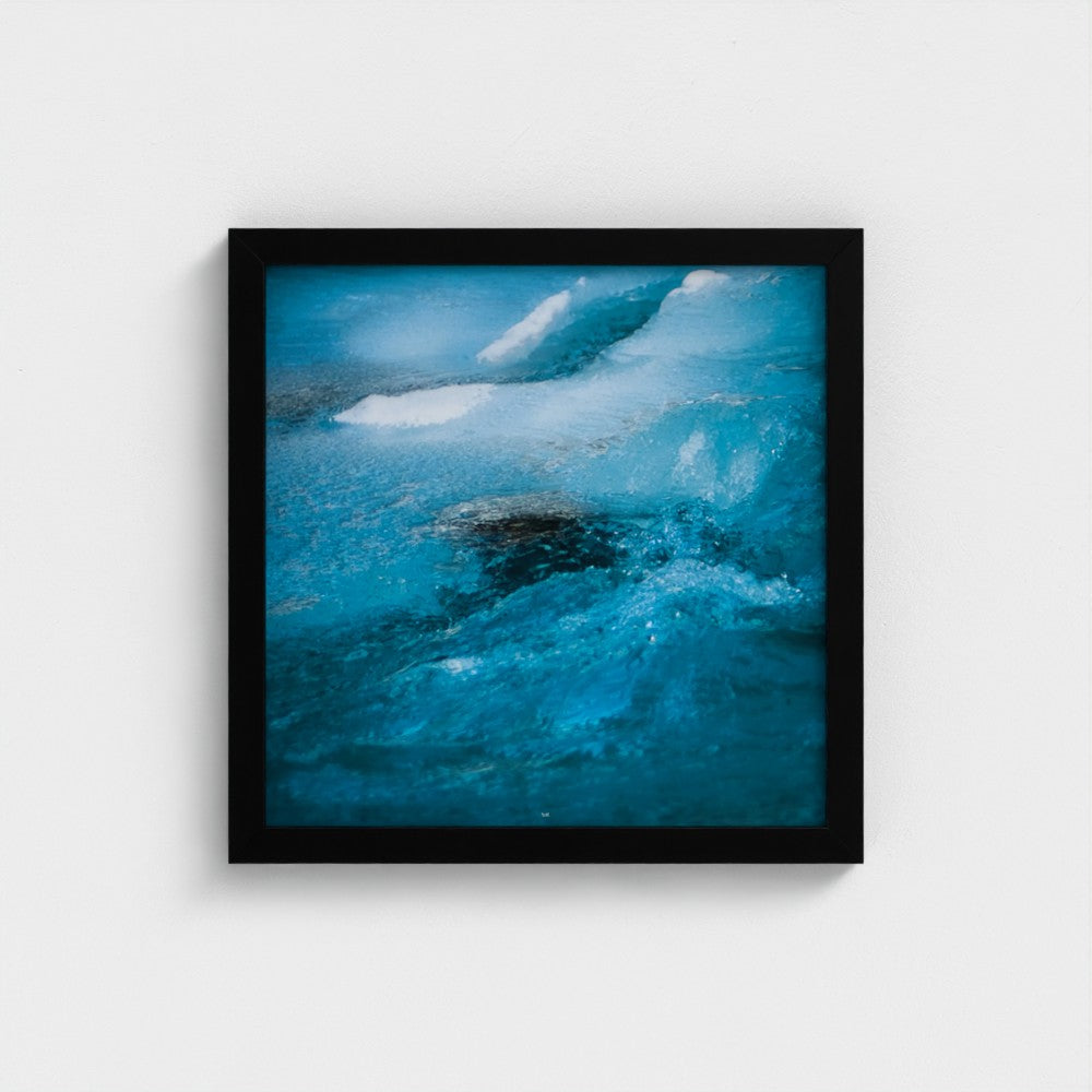 A framed square art print featuring a close-up photograph of vibrant blue ice or water with white highlights, enclosed in a simple black frame against a light gray wall.