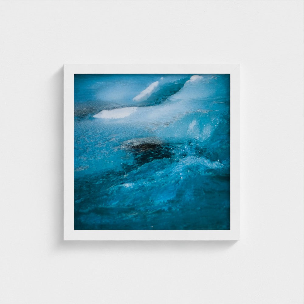 A square framed abstract art print depicting vibrant blue and turquoise water waves, mounted on a clean white wall.