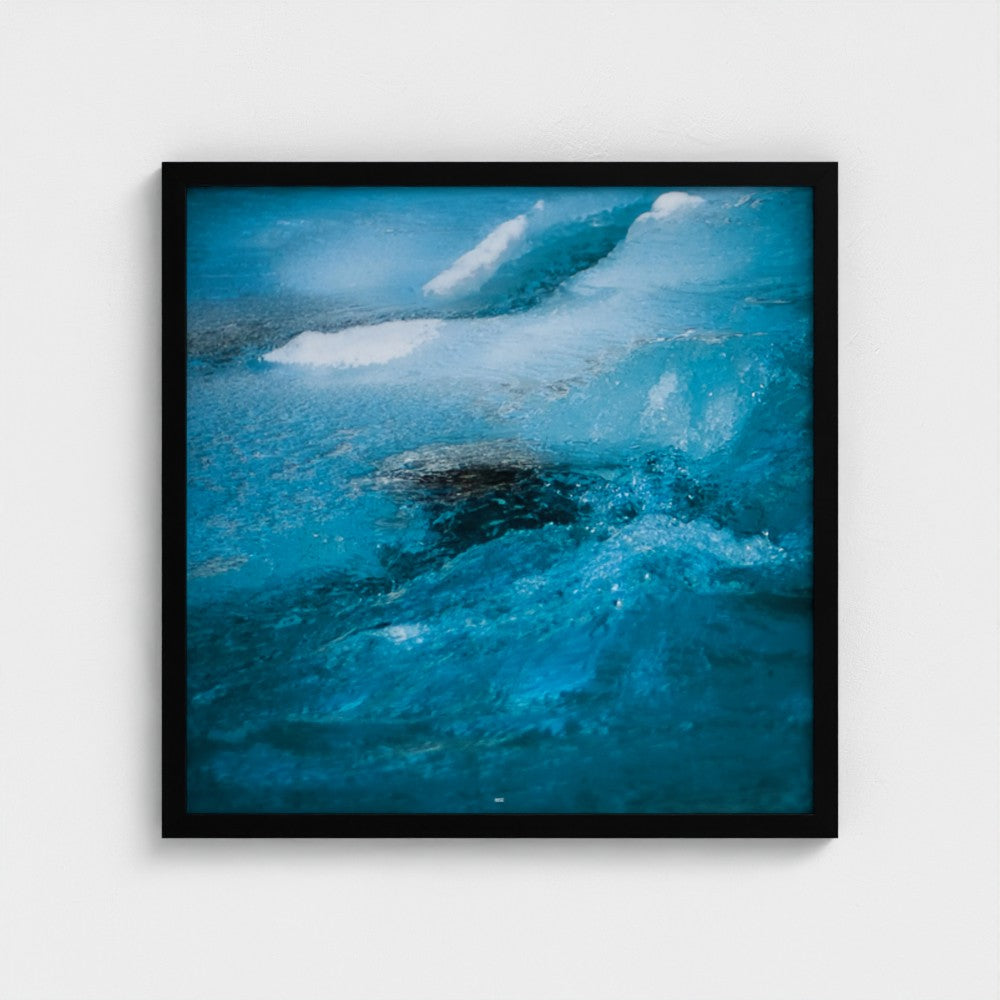 A square framed art print depicting a close-up abstract view of turquoise ocean waves with white foam, set in a simple black frame against a light gray wall.