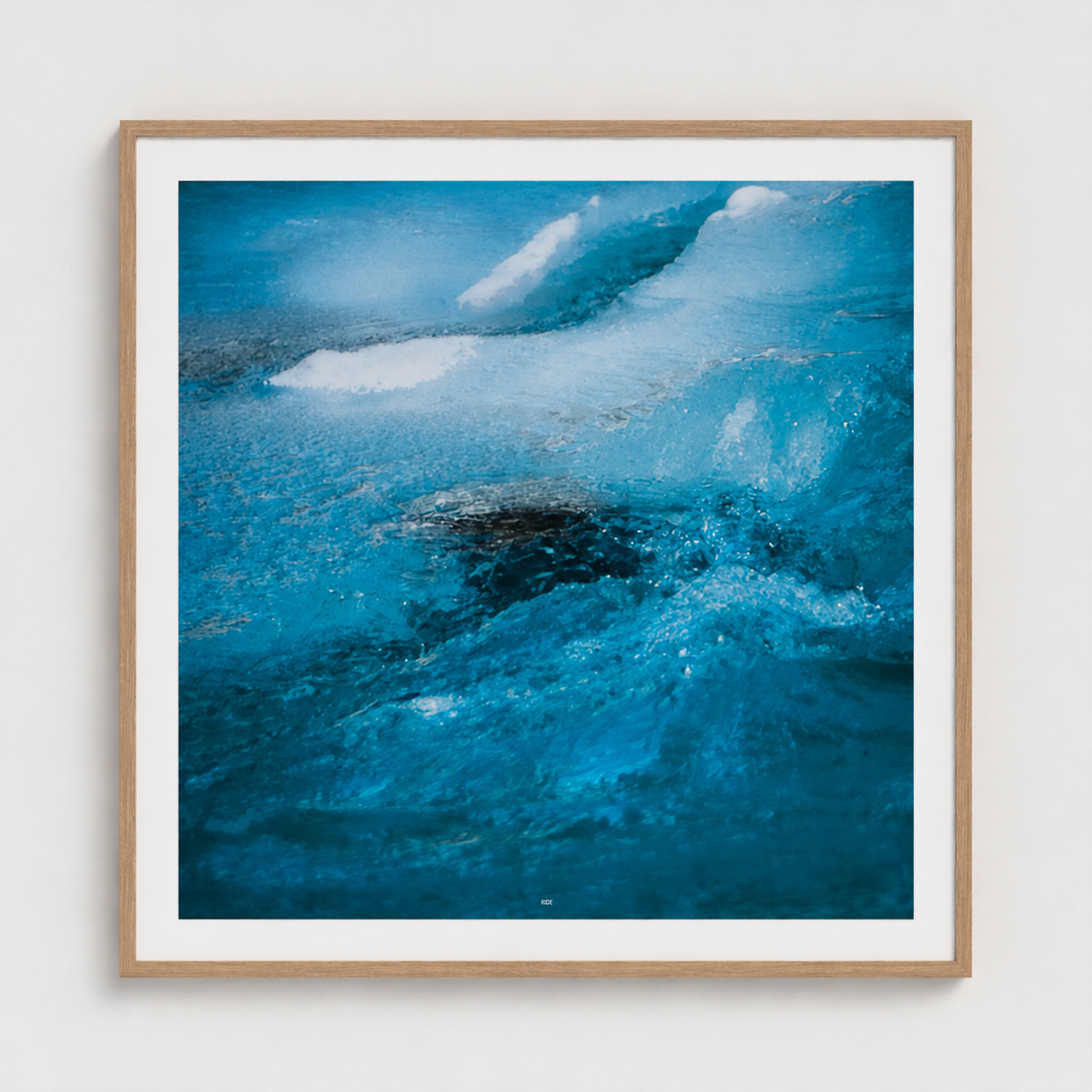 A square framed art print depicting a close-up of vibrant blue ocean waves with white foam, set in a light natural wood frame with a white mat against a neutral light gray wall.