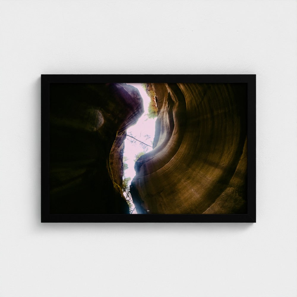 A framed photographic print of a slot canyon looking up at light filtering through, displayed on a clean white wall.