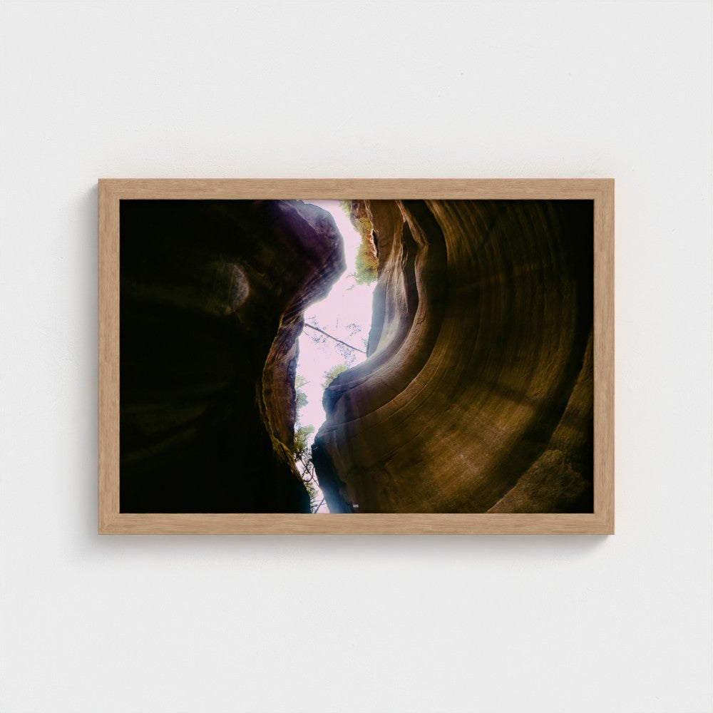 A framed landscape photograph of a slot canyon looking up at light filtering through, presented in a light natural wood frame against a white wall.