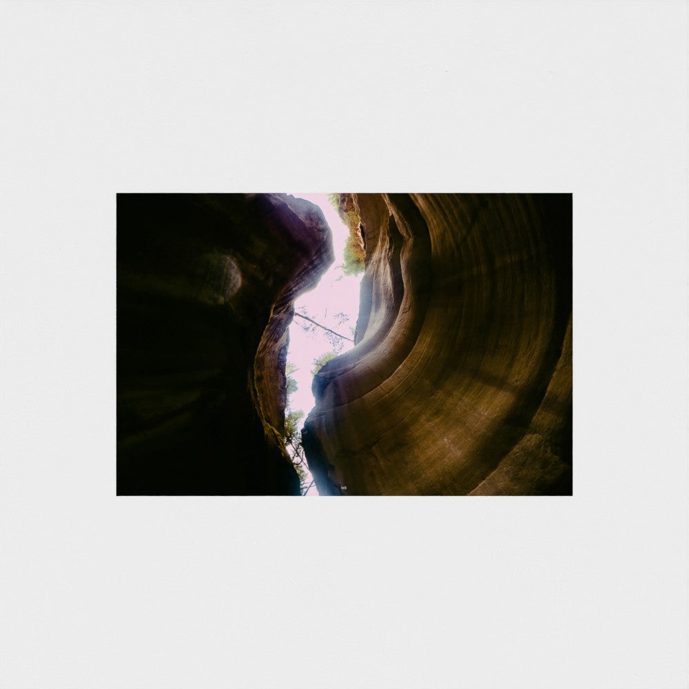 A rectangular art print depicting a dramatic low-angle view inside a slot canyon, looking up towards a bright opening of light. The print is centered on a solid light gray background.