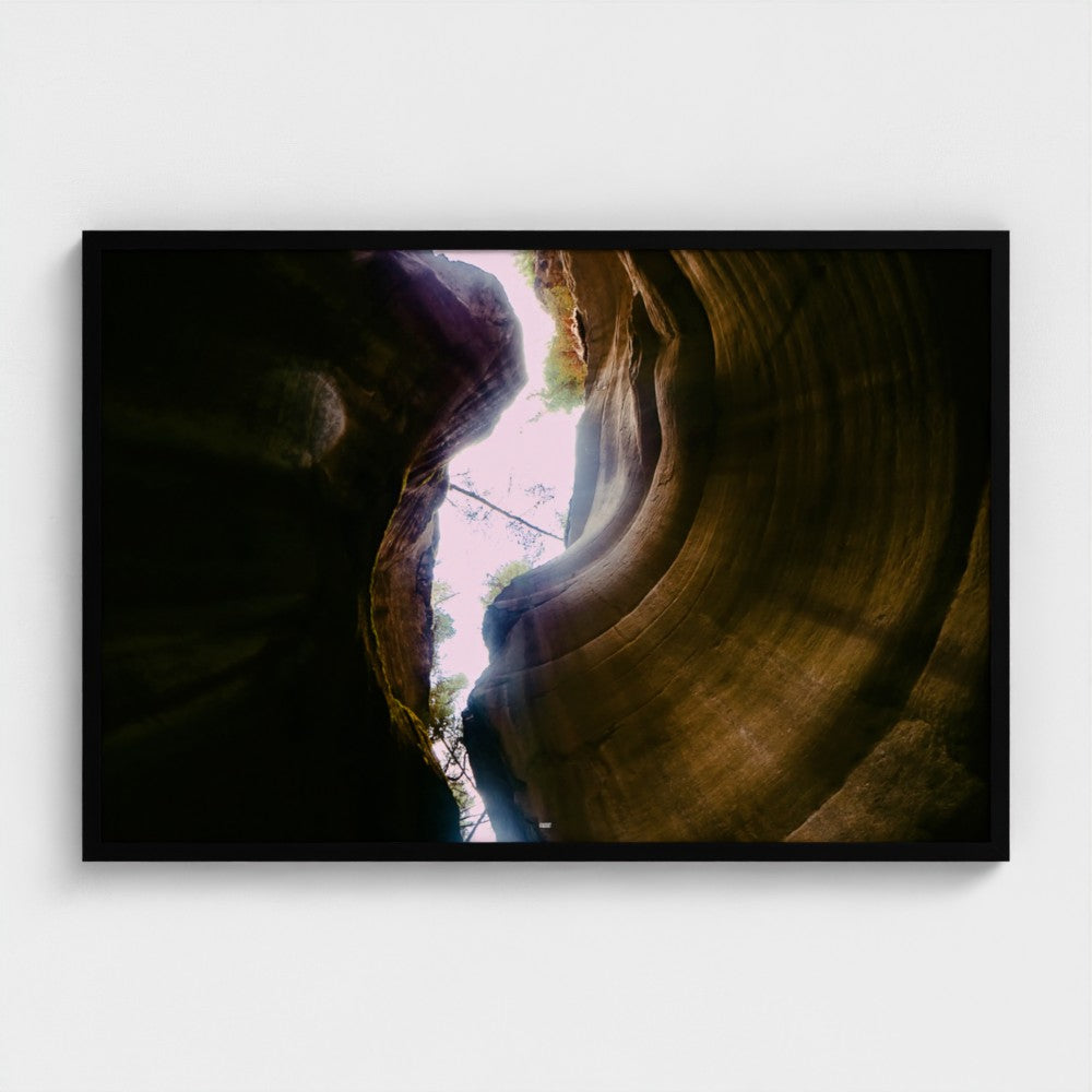 A framed landscape art print depicting a slot canyon with light rays shining through, mounted on a light gray wall.