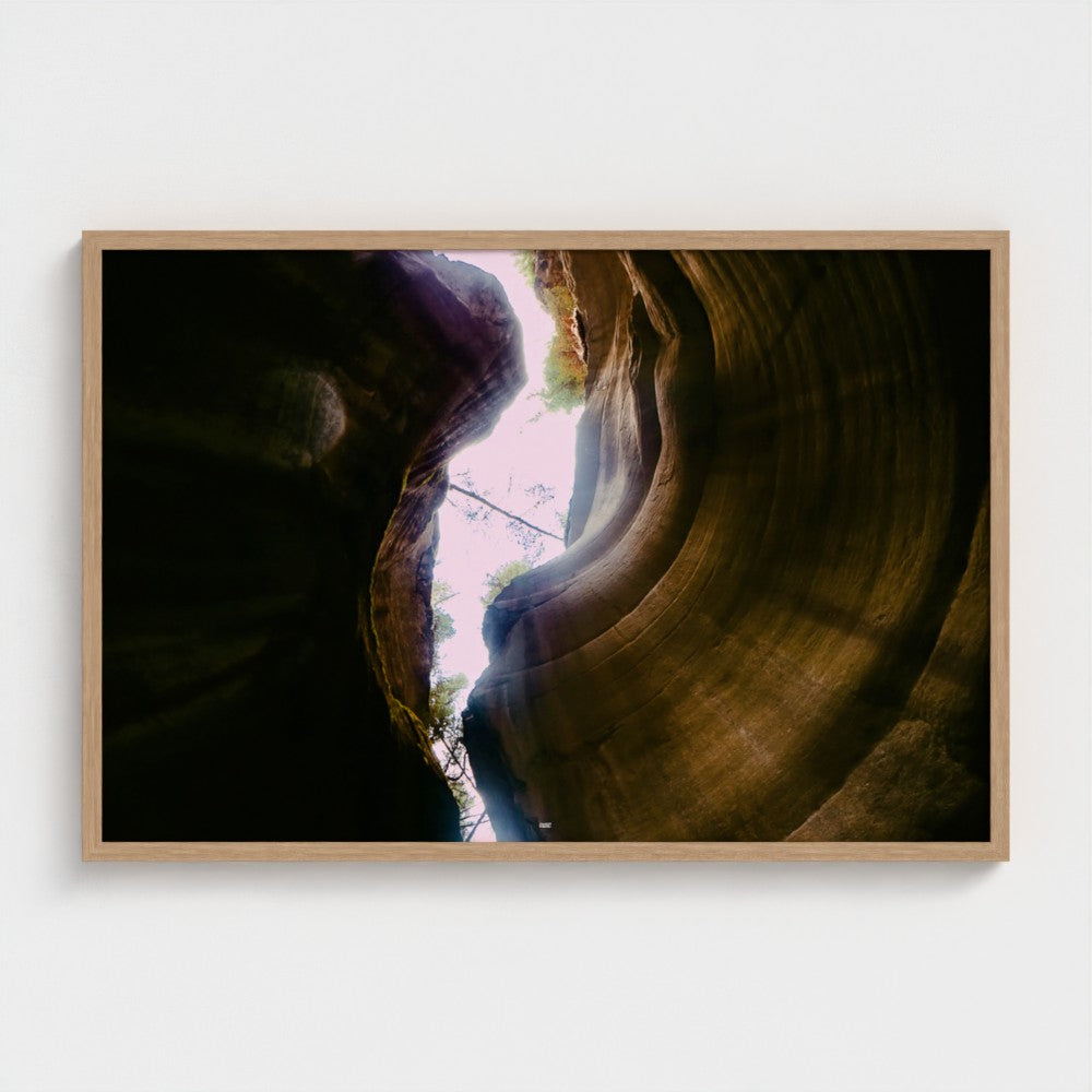 A framed photographic print of a slot canyon looking up at the opening, featuring warm brown and black rock curves with a bright light source above, mounted on a light gray wall.