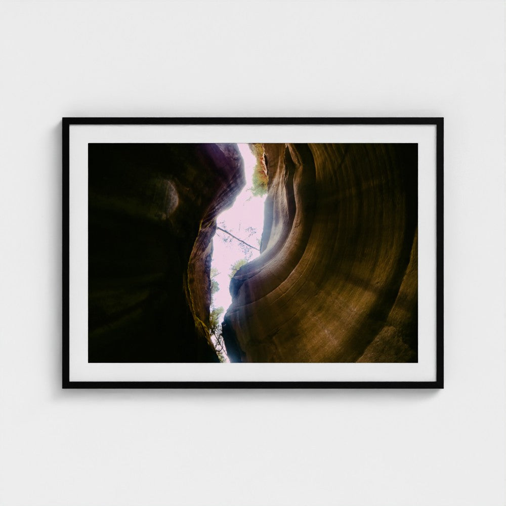A framed art print depicting a dramatic canyon scene with light streaming into a deep gorge, presented in a black frame with a white mat against a light gray wall.