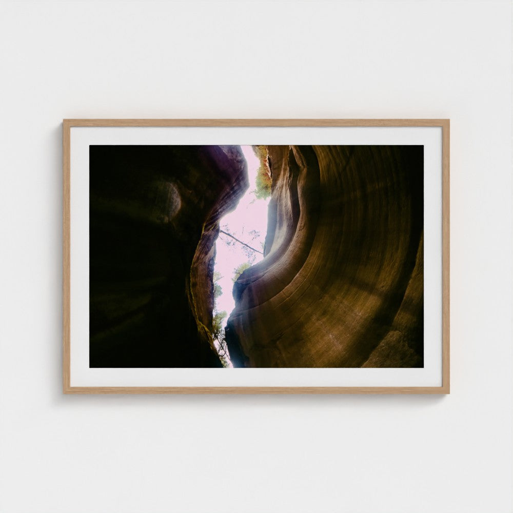 A framed photographic print of a canyon interior with a bright opening of light, displayed on a clean white wall.