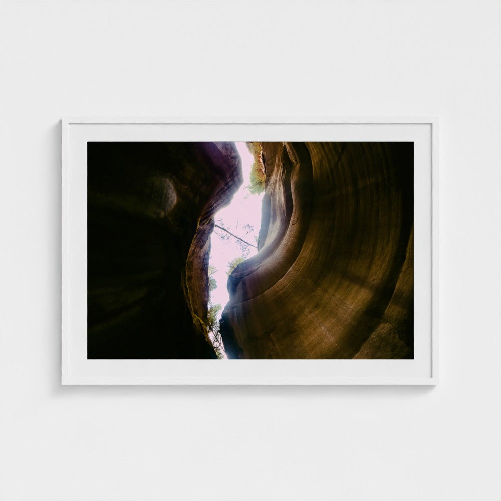 A framed photographic print of a canyon interior with a bright opening of light, displayed on a clean white wall.