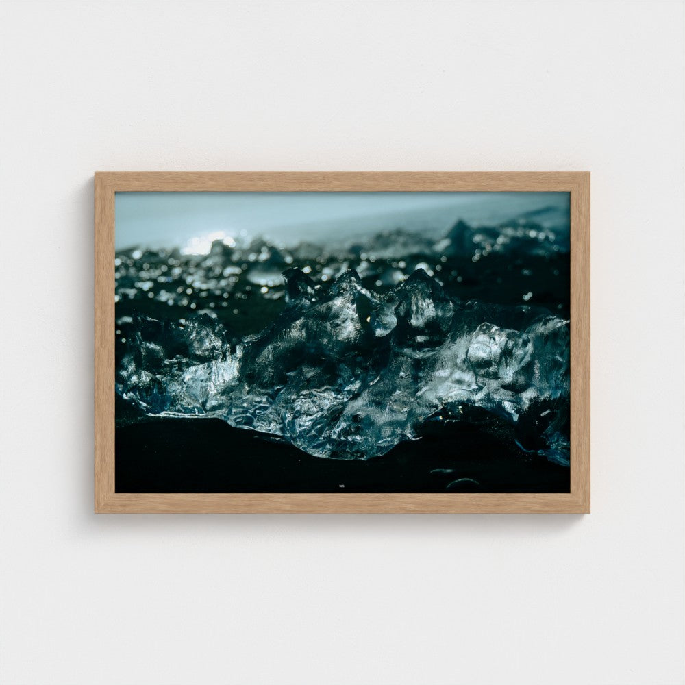 A framed photographic print of ice cubes on a black sand beach, enclosed in a light natural wood frame and mounted on a plain white wall.