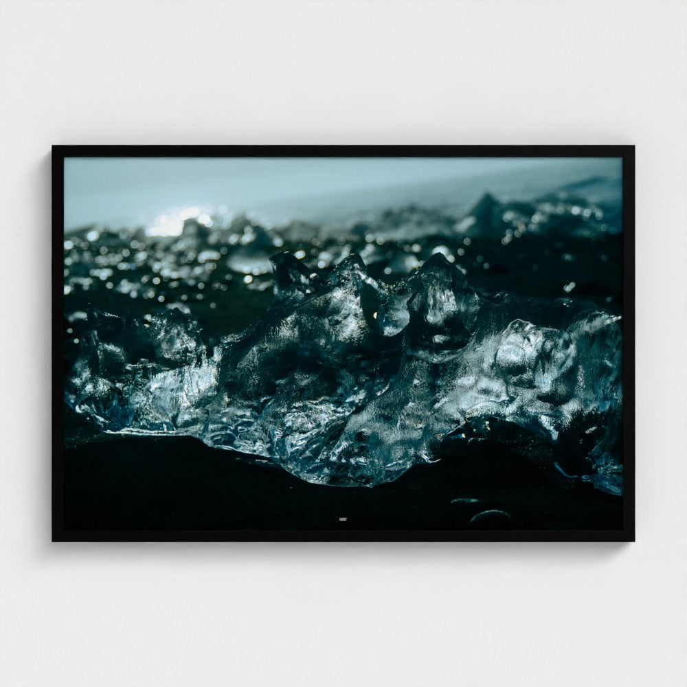A framed photographic print of dark, icy blue-black icebergs on a black sand beach, presented in a black frame against a light gray wall.