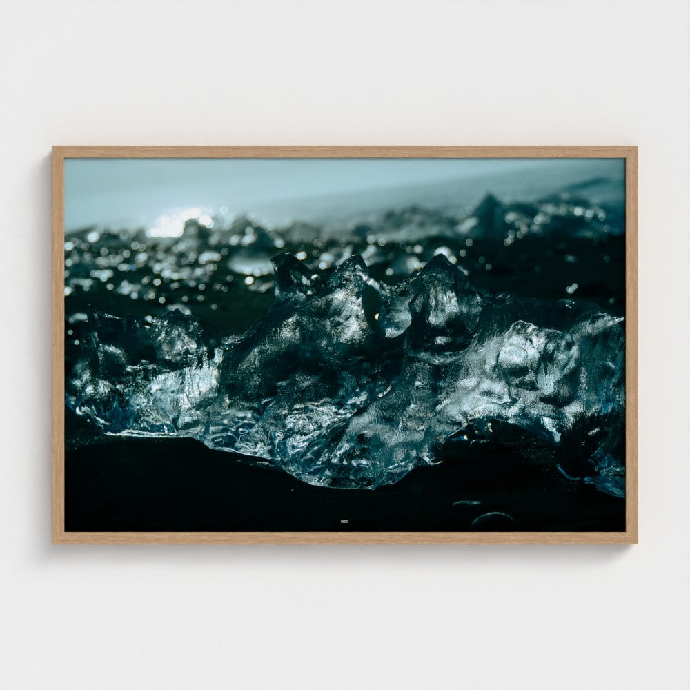 A framed photographic print of dark blue-black ice formations on a black sand beach, mounted on a light gray wall.