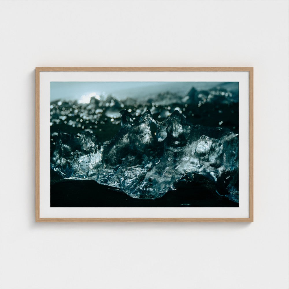 A framed photographic art print depicting a close-up of dark, textured ice or black sand with blue-gray highlights, presented in a light wood frame with a white mat against a clean white wall.