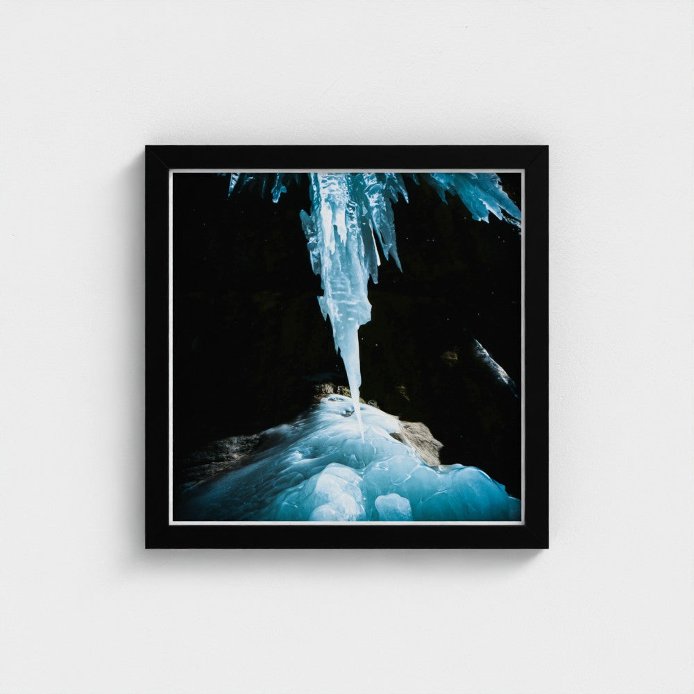 A framed square wall art print showing a dramatic icy blue icicle hanging over frozen blue ice in a dark cave, displayed against a light gray wall.
