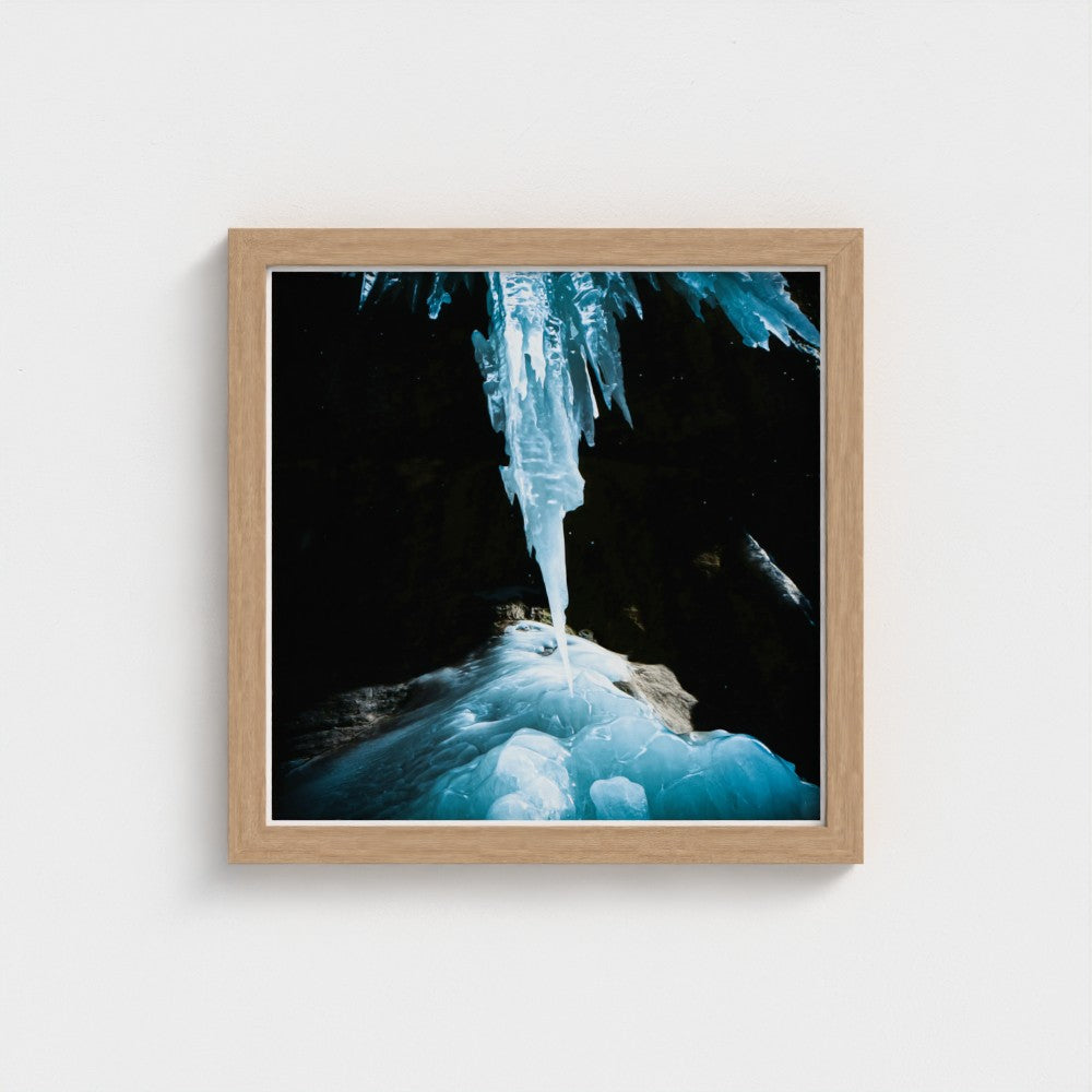 Framed wall art featuring a dramatic photograph of a glowing blue ice formation hanging in a dark cave, presented in a light natural wood frame against a white wall.