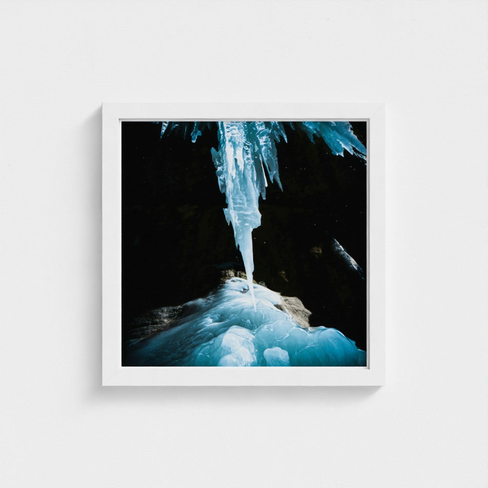 A framed square art print featuring a photograph of a glowing blue ice formation hanging in a dark cave, presented in a white frame against a light gray wall.