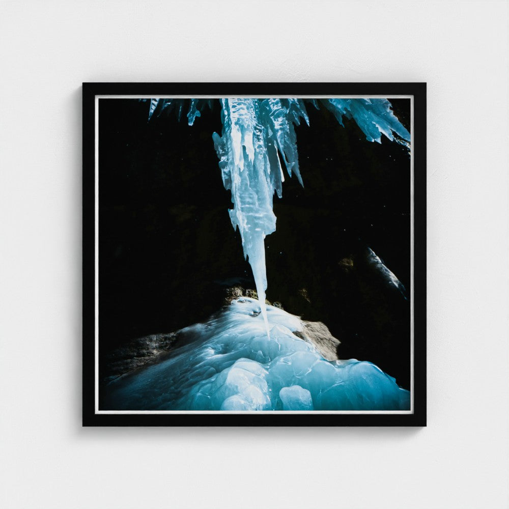 Framed wall art featuring a dramatic close-up photograph of a glowing blue ice formation hanging from a dark cave ceiling, presented in a black frame with a white mat against a light gray background.
