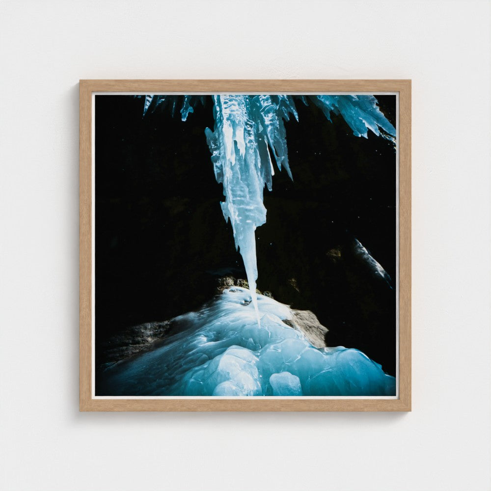 Framed wall art featuring a dramatic photograph of a glowing blue ice column hanging from a cave ceiling, set in a light natural wood frame against a white wall.