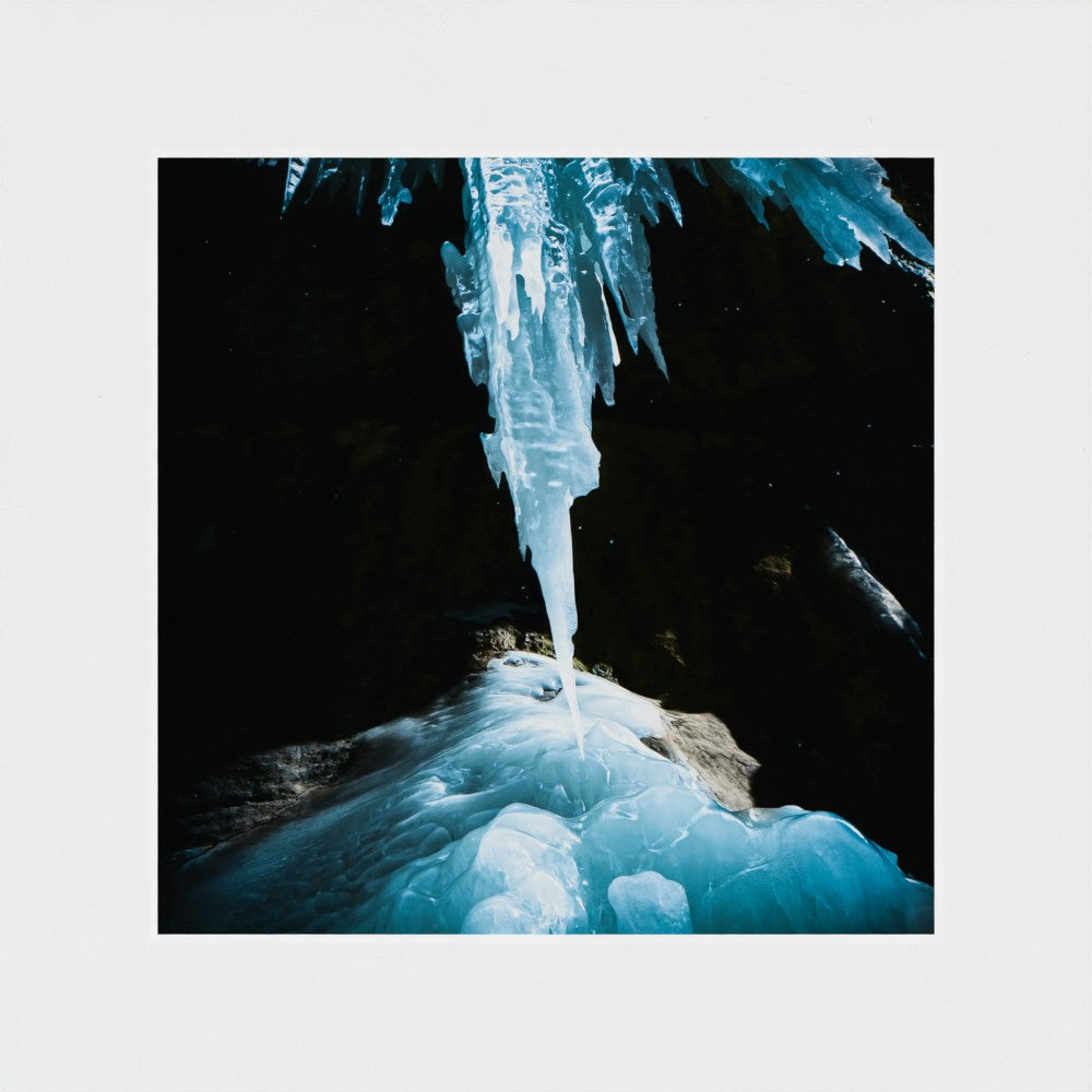 A square photographic print of a dramatic ice cave scene, featuring a large icicle hanging from a dark cave ceiling and blue frozen rock below, centered on a light gray background.