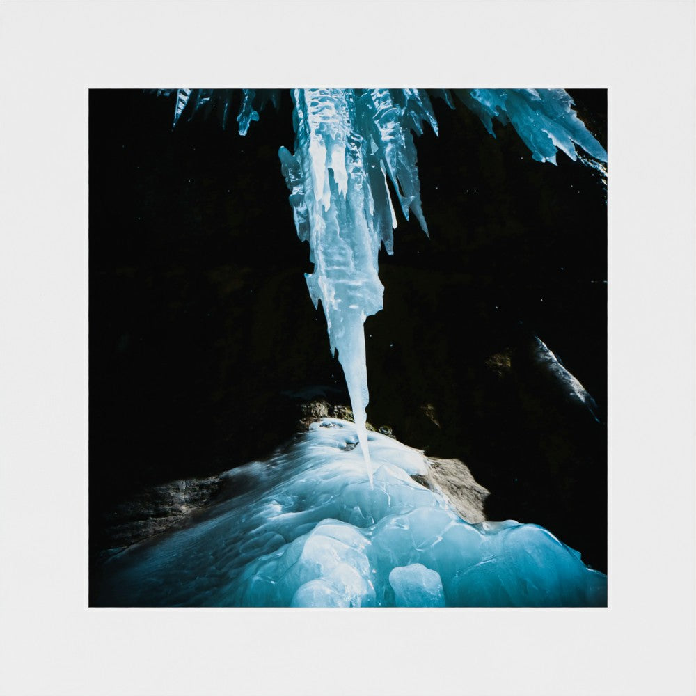 A square photographic print of a dramatic ice formation hanging from a dark cave ceiling, with bright blue and white frozen structures contrasting against a black background, centered on a light gray border.