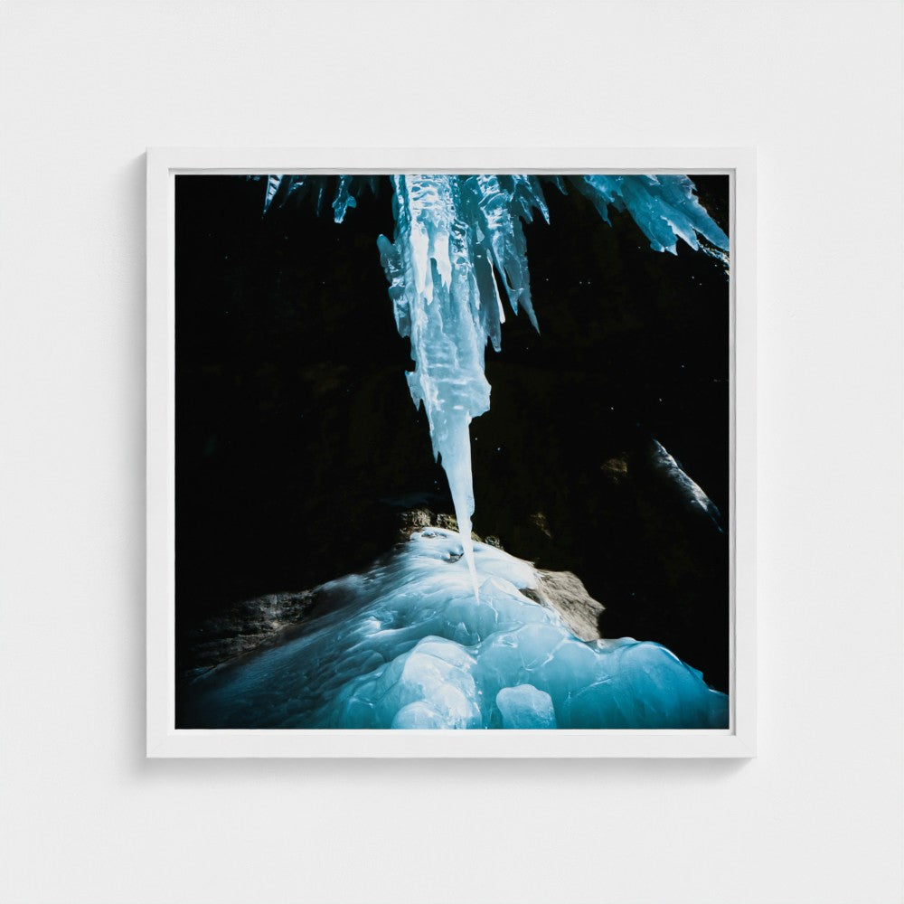 A framed square art print featuring a dramatic photograph of a glowing blue ice icicle hanging over frozen rock in a dark cave, presented in a white frame against a light gray wall.