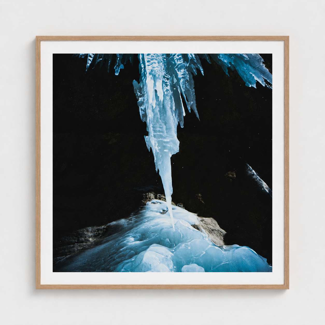 A framed photographic art print depicting a dramatic ice cave scene with a large icicle hanging from a dark cave ceiling and blue ice formations below, presented in a light wood frame with a white mat against a light gray wall.