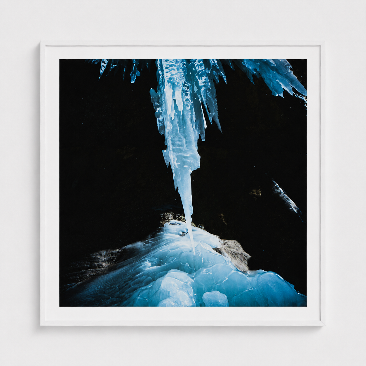 A framed square art print depicting a dramatic frozen icicle hanging over blue ice formations against a dark background, presented in a white frame on a light gray wall.