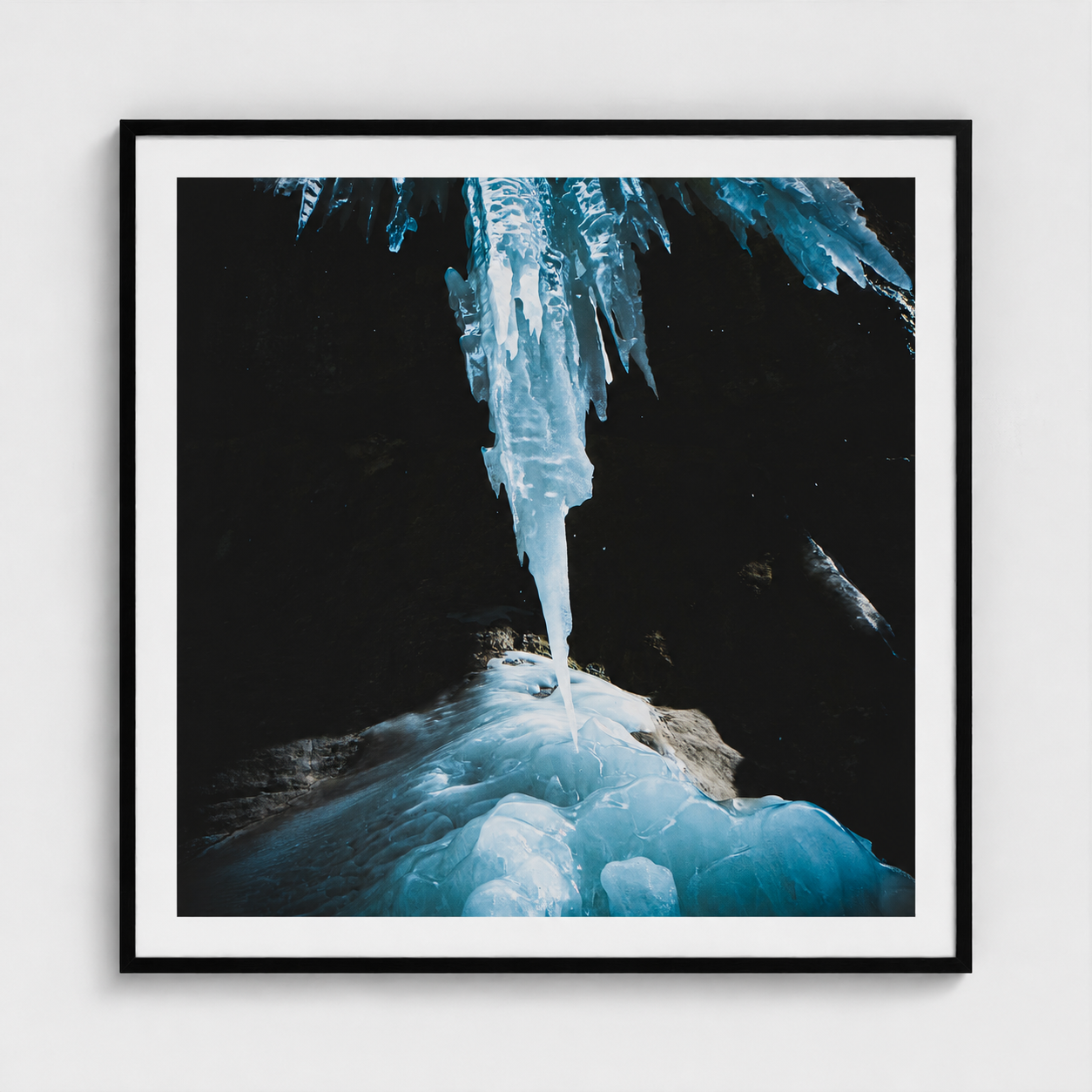 A framed photographic art print depicting a dramatic icicle hanging from a dark cave ceiling over a frozen blue and white ice formation. The print is presented with a white mat and a thin black frame against a light gray wall.