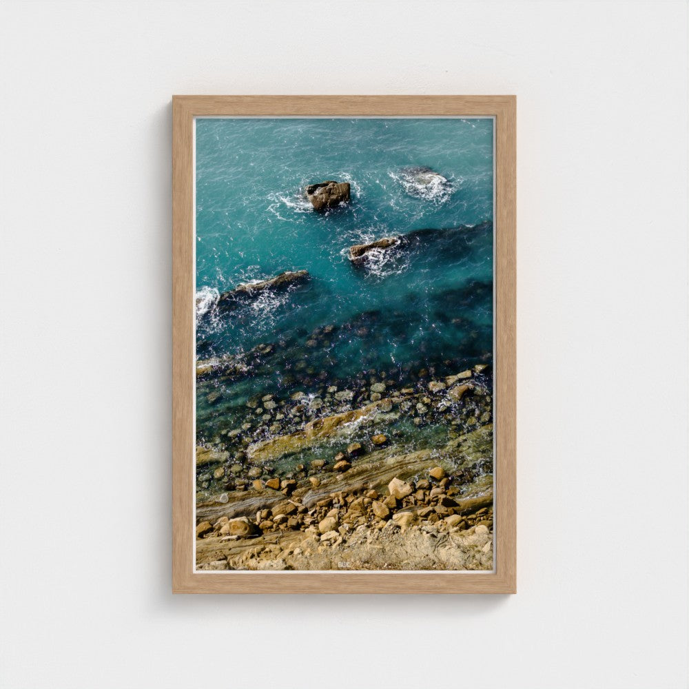 Framed artwork of a coastal scene with rocks and water on a white wall