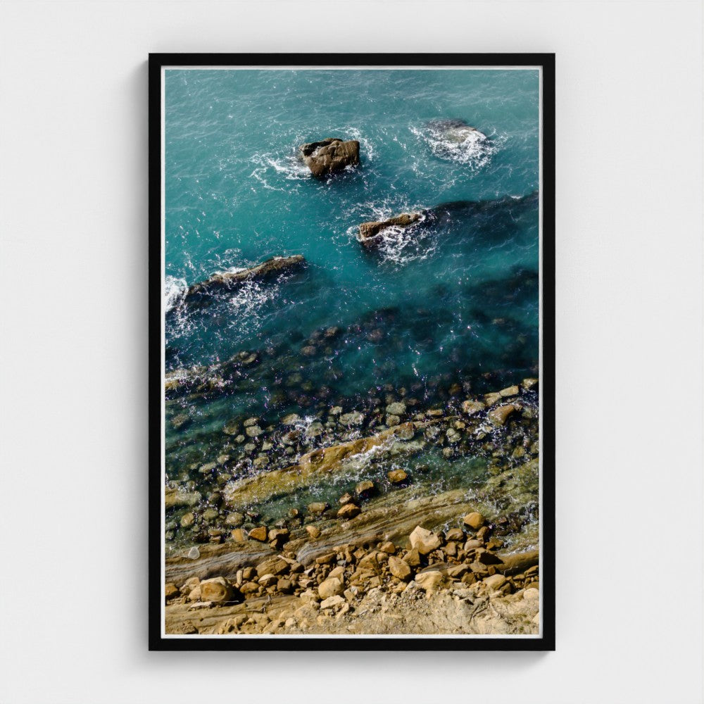 Framed artwork of a coastal scene with rocks and blue water.