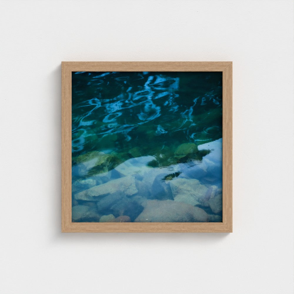 A square framed art print depicting a close-up of clear blue and green water over pale rocks, set in a light natural wood frame against a white wall.