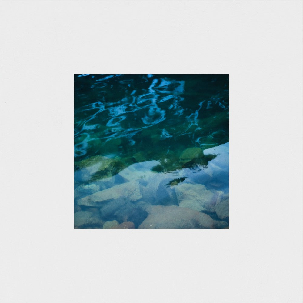 A square photographic print depicting clear blue and green water over pale rocks, centered on a light gray background.