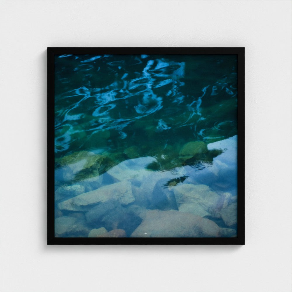 A square framed art print depicting a close-up of clear turquoise water over pale rocks, with a black frame and a subtle drop shadow on a light gray background.