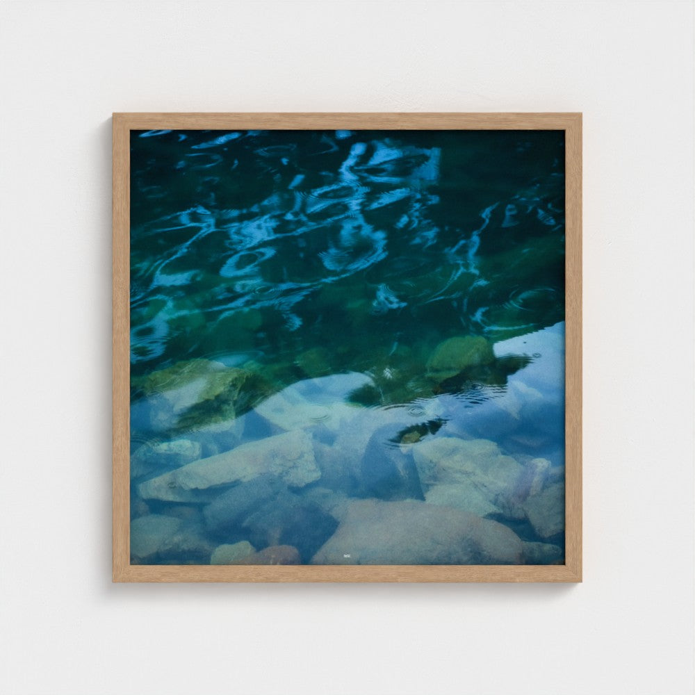 A square framed art print depicting clear blue and green water over light-colored rocks, set in a light wood frame against a plain white wall.