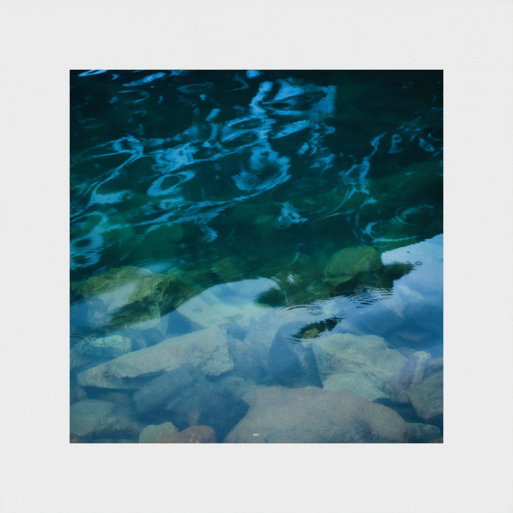 A square photographic print depicting clear blue and green water over smooth rocks, centered on a light gray background.