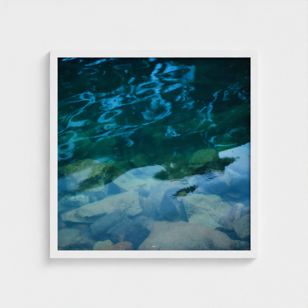 A framed square art print depicting a close-up photograph of clear turquoise water over smooth rocks, displayed against a light gray wall.