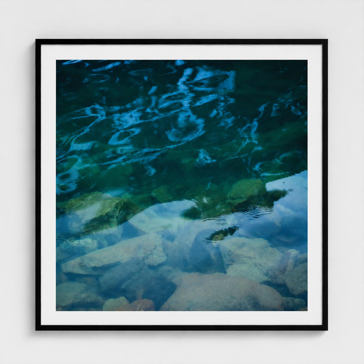 Framed square wall art featuring a close-up photograph of clear blue and green water over rocks, displayed against a light gray wall.