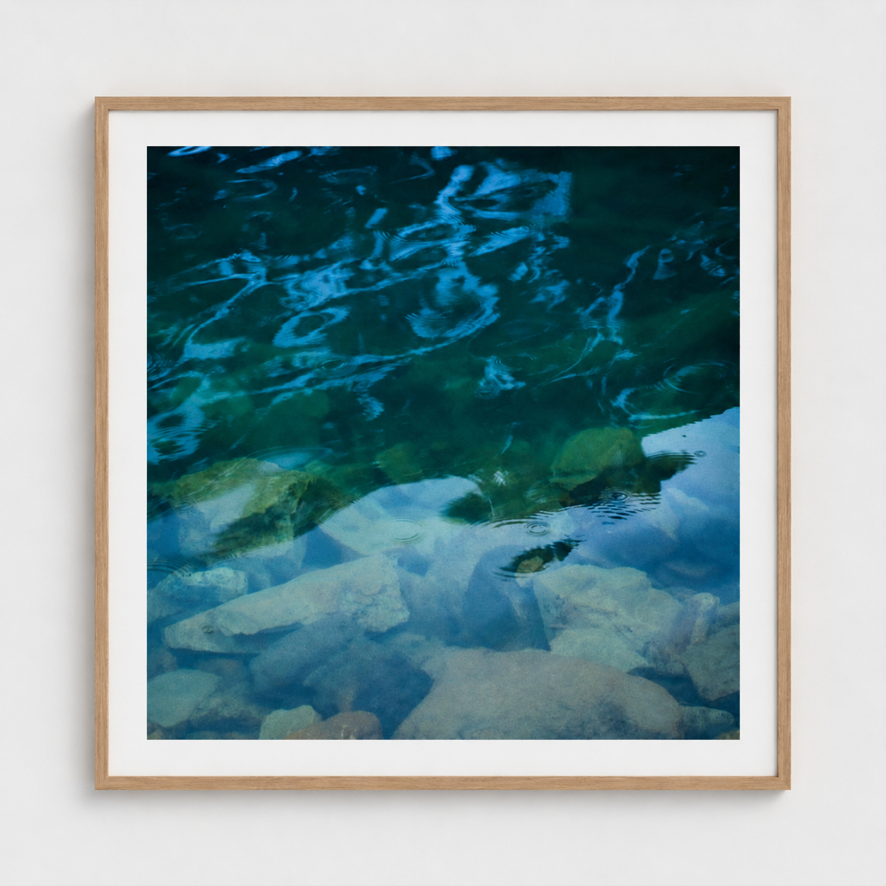 A square framed art print depicting a close-up photograph of clear blue and green water over rocks, set in a light wood frame with a white mat against a light gray wall.